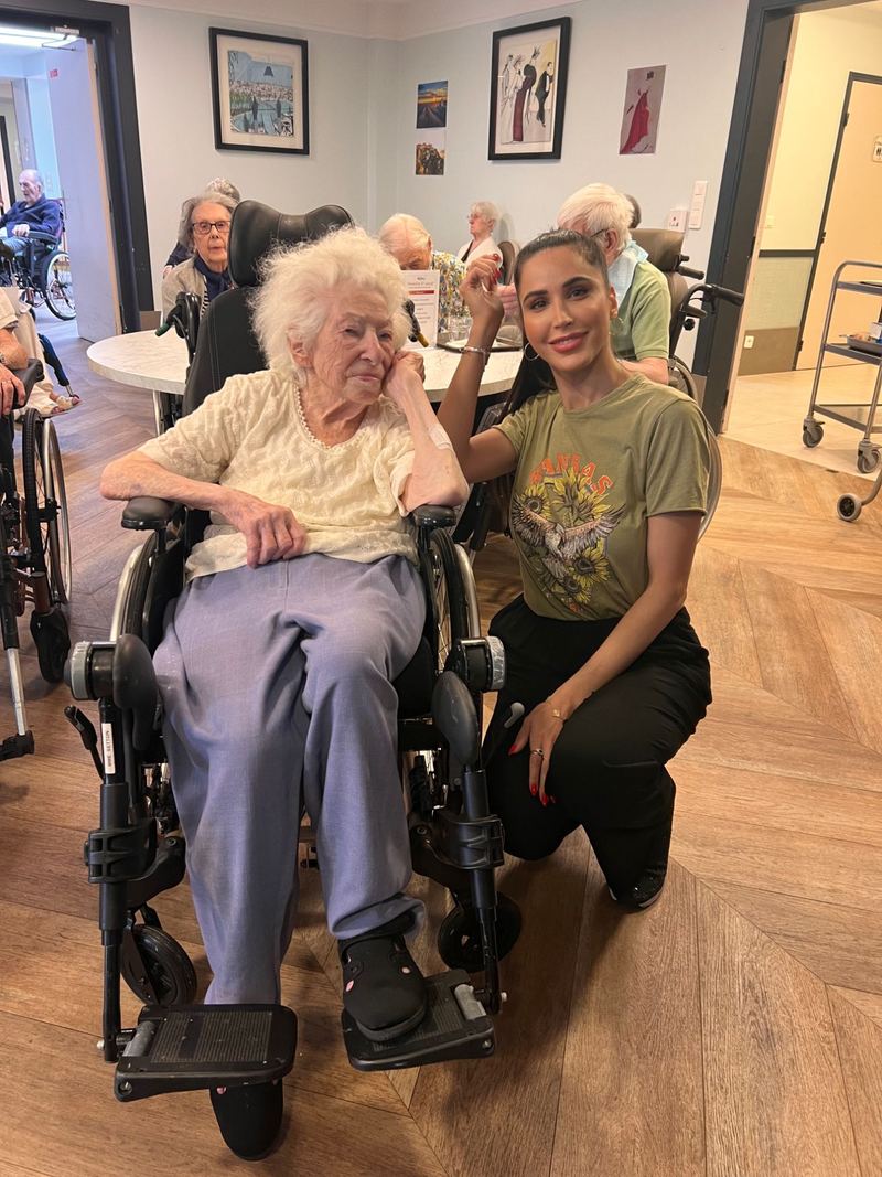 Somayeh accroupie à côté d’une résidente âgée en fauteuil roulant, dans une maison de retraite
